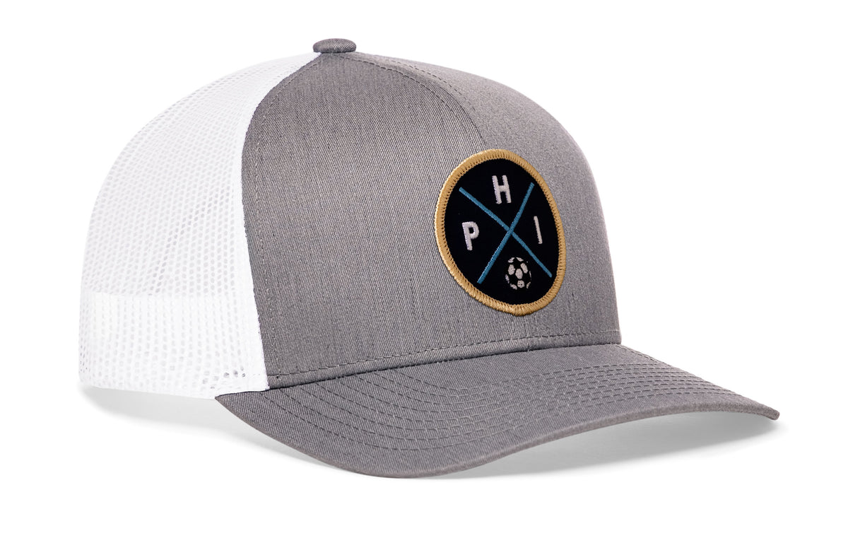 gray-white trucker hat viewed from the right side, showcasing a bold navy and gold embroidered X patch prominently displayed on the front