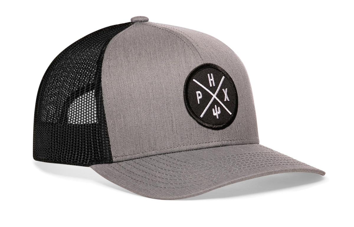 gray-black trucker hat viewed from the right side, showcasing a bold black and white embroidered X patch prominently displayed on the front