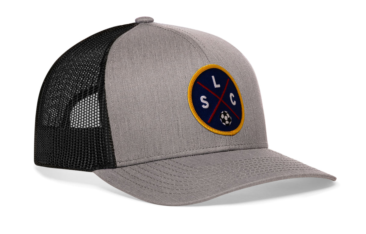 gray-black trucker hat viewed from the right side, showcasing a bold deep red and blue embroidered X patch prominently displayed on the front