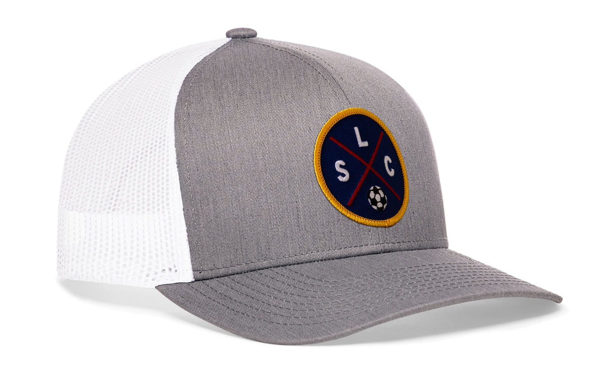 gray-white trucker hat viewed from the right side, showcasing a bold deep red and blue embroidered X patch prominently displayed on the front