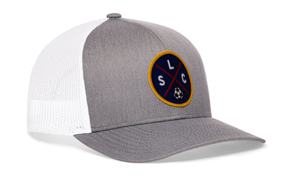 gray-white trucker hat viewed from the right side, showcasing a bold deep red and blue embroidered X patch prominently displayed on the front