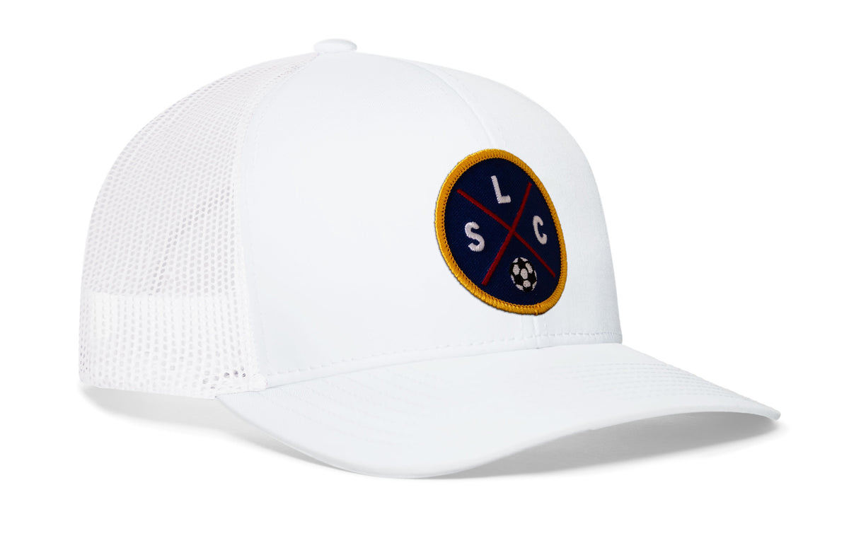 white trucker hat viewed from the right side, showcasing a bold deep red and blue embroidered X patch prominently displayed on the front