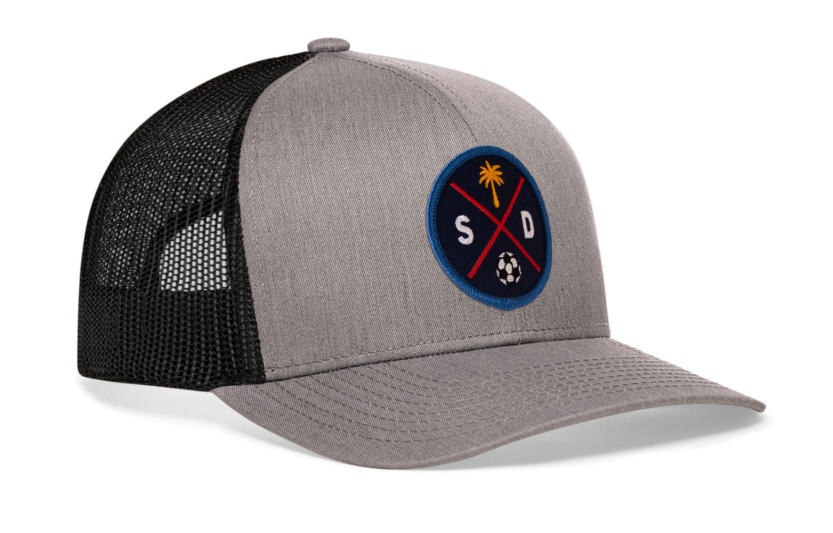 gray-black trucker hat viewed from the right side, showcasing a bold navy and orange embroidered X patch prominently displayed on the front