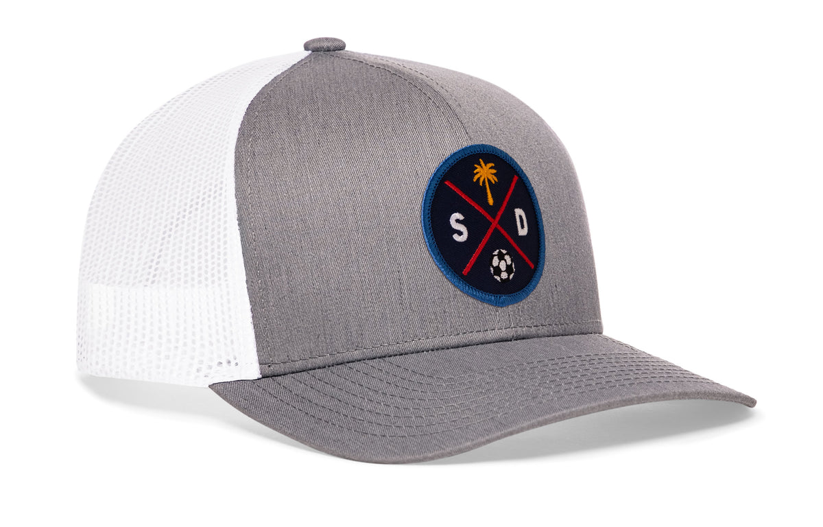 gray-white trucker hat viewed from the right side, showcasing a bold navy and orange embroidered X patch prominently displayed on the front