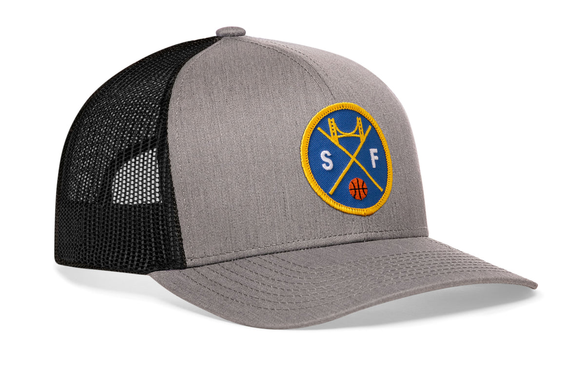 gray-black trucker hat viewed from the right side, showcasing a bold royal blue and gold embroidered X patch prominently displayed on the front
