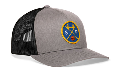 gray-black trucker hat viewed from the right side, showcasing a bold royal blue and gold embroidered X patch prominently displayed on the front