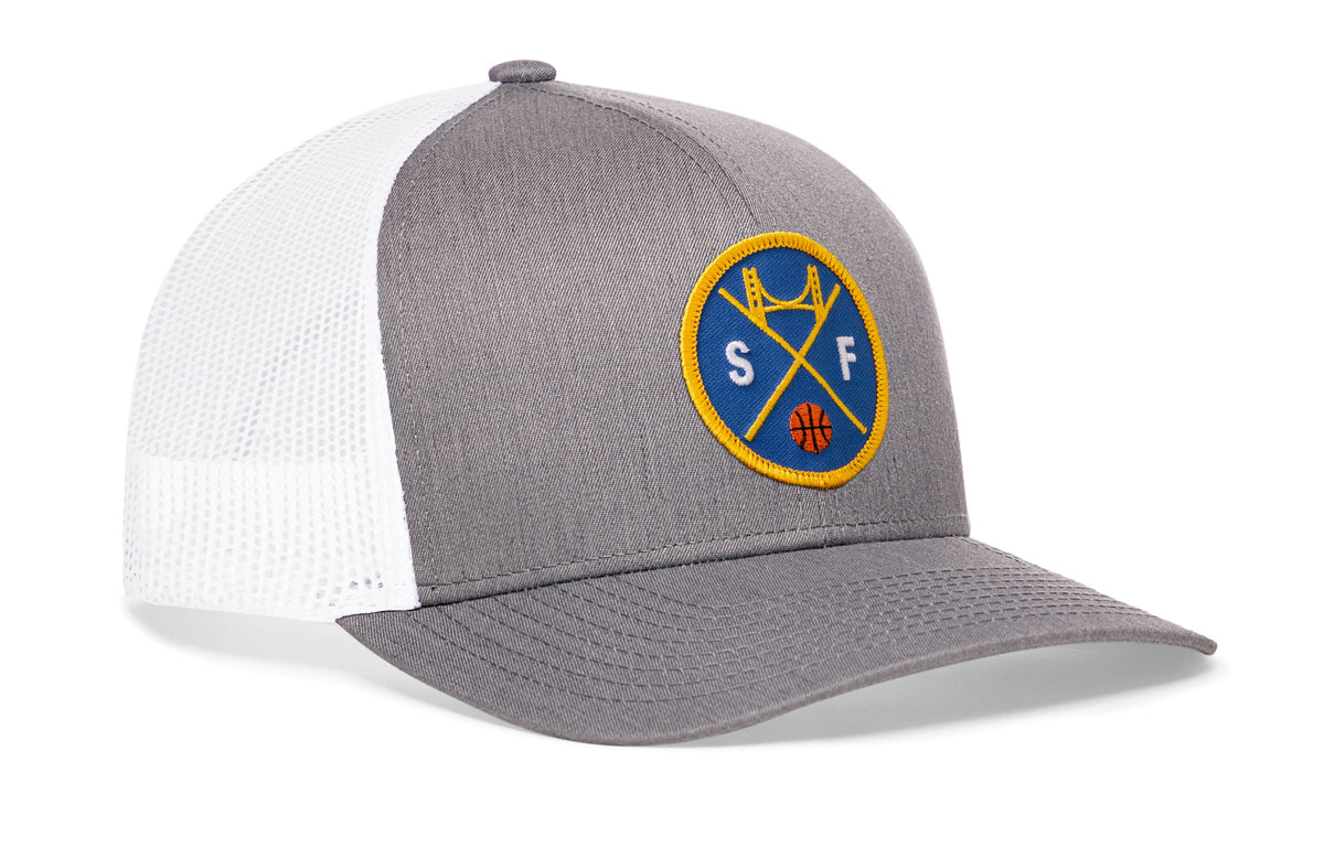 gray-white trucker hat viewed from the right side, showcasing a bold royal blue and gold embroidered X patch prominently displayed on the front