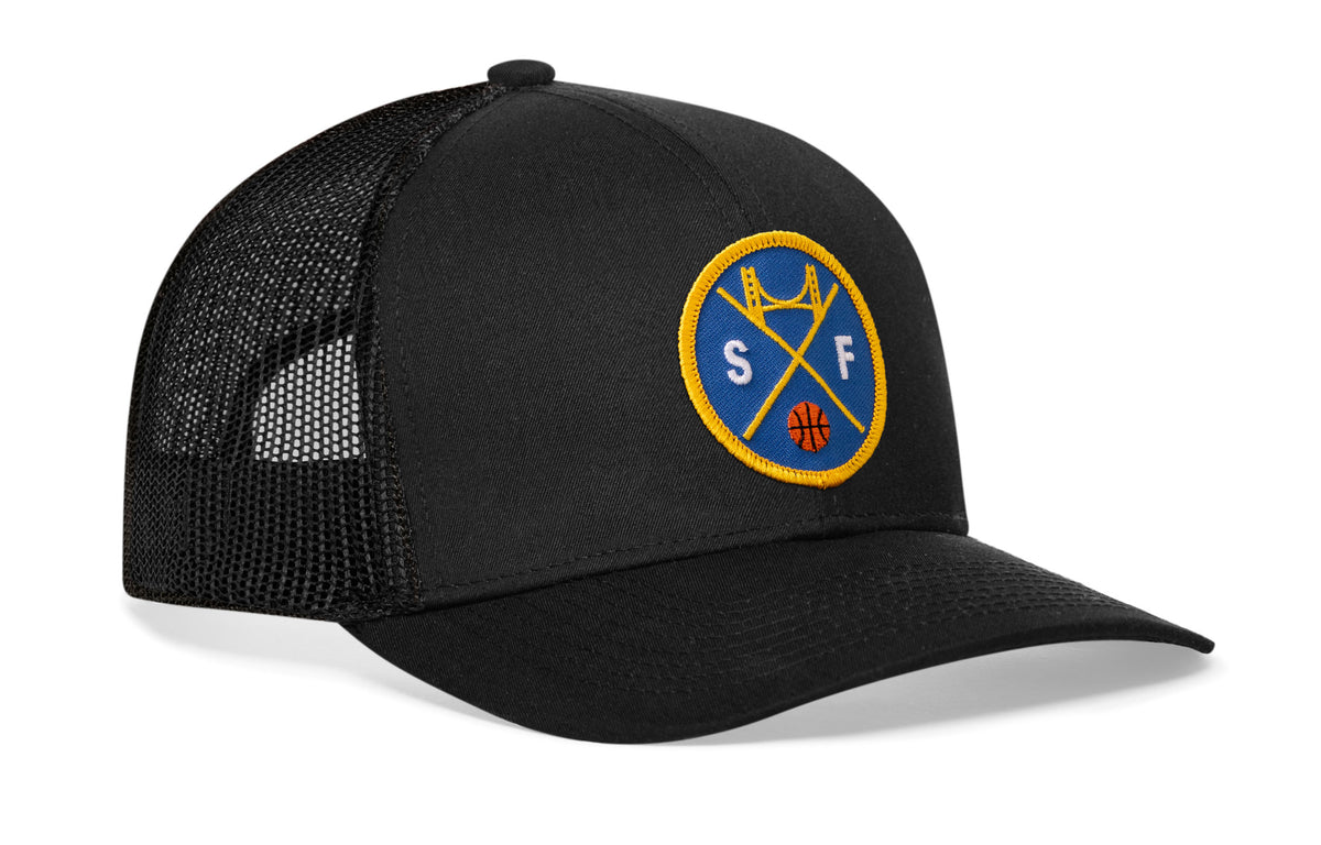 black trucker hat viewed from the right side, showcasing a bold royal blue and gold embroidered X patch prominently displayed on the front