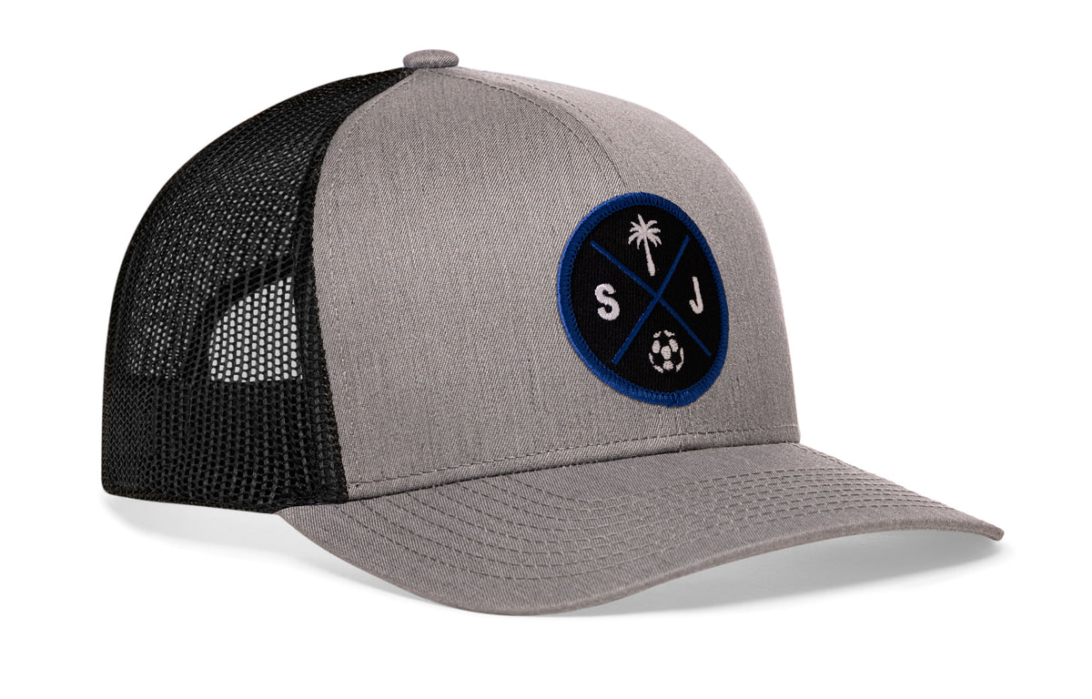 gray-black trucker hat viewed from the right side, showcasing a bold black and blue embroidered X patch prominently displayed on the front