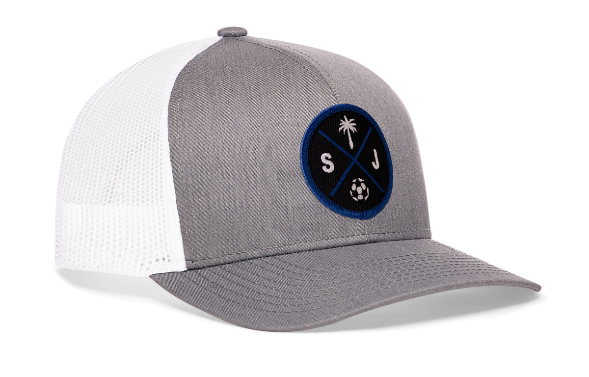 gray-white trucker hat viewed from the right side, showcasing a bold black and blue embroidered X patch prominently displayed on the front