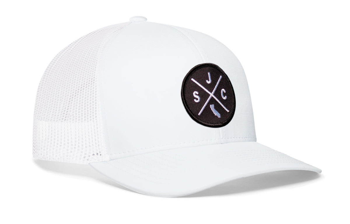 white trucker hat viewed from the right side, showcasing a bold black and white embroidered X patch prominently displayed on the front