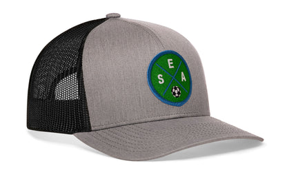 gray-black trucker hat viewed from the right side, showcasing a bold green and blue embroidered X patch prominently displayed on the front