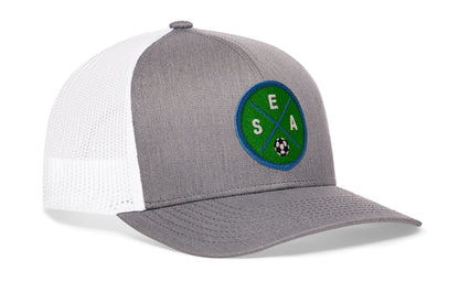 gray-white trucker hat viewed from the right side, showcasing a bold green and blue embroidered X patch prominently displayed on the front