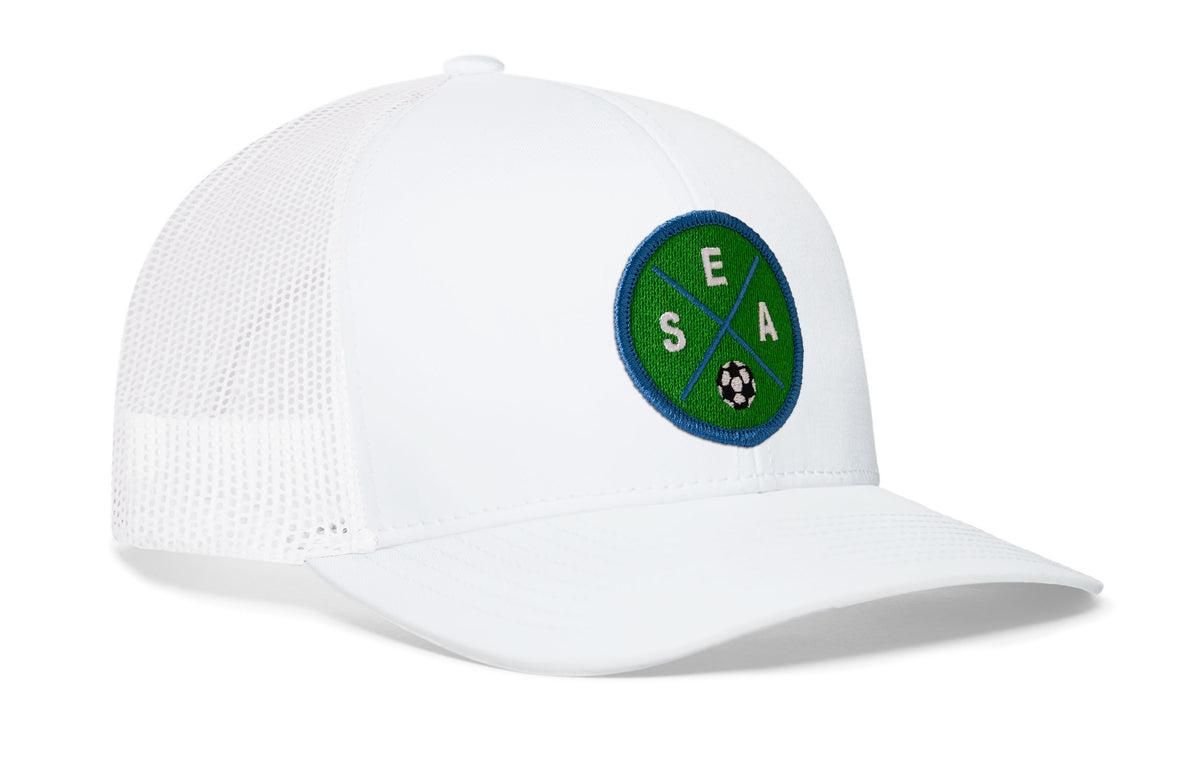 white trucker hat viewed from the right side, showcasing a bold green and blue embroidered X patch prominently displayed on the front