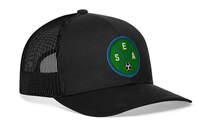 black trucker hat viewed from the right side, showcasing a bold green and blue embroidered X patch prominently displayed on the front