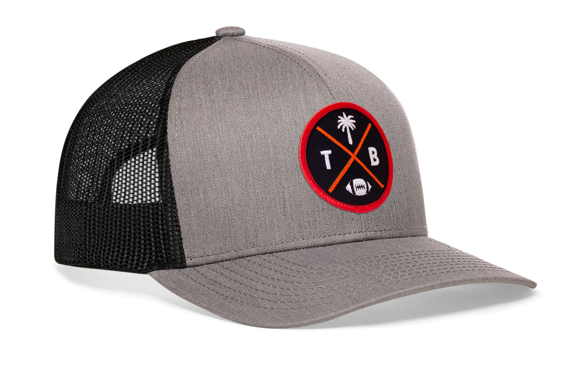 gray-black trucker hat viewed from the right side, showcasing a bold red, orange and black embroidered X patch prominently displayed on the front