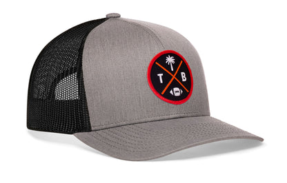gray-black trucker hat viewed from the right side, showcasing a bold red, orange and black embroidered X patch prominently displayed on the front