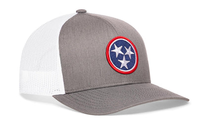 gray-white trucker hat viewed from the right side, showcasing a bold red, white and blue embroidered patch prominently displayed on the front