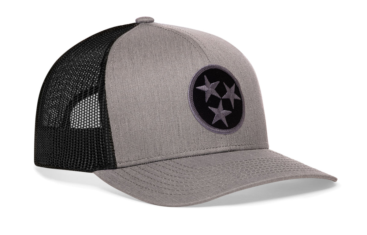 gray-black trucker hat viewed from the right side, showcasing a bold black and gray embroidered flag patch prominently displayed on the front
