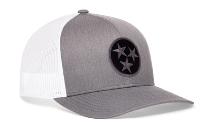 gray-white trucker hat viewed from the right side, showcasing a bold black and gray embroidered flag patch prominently displayed on the front
