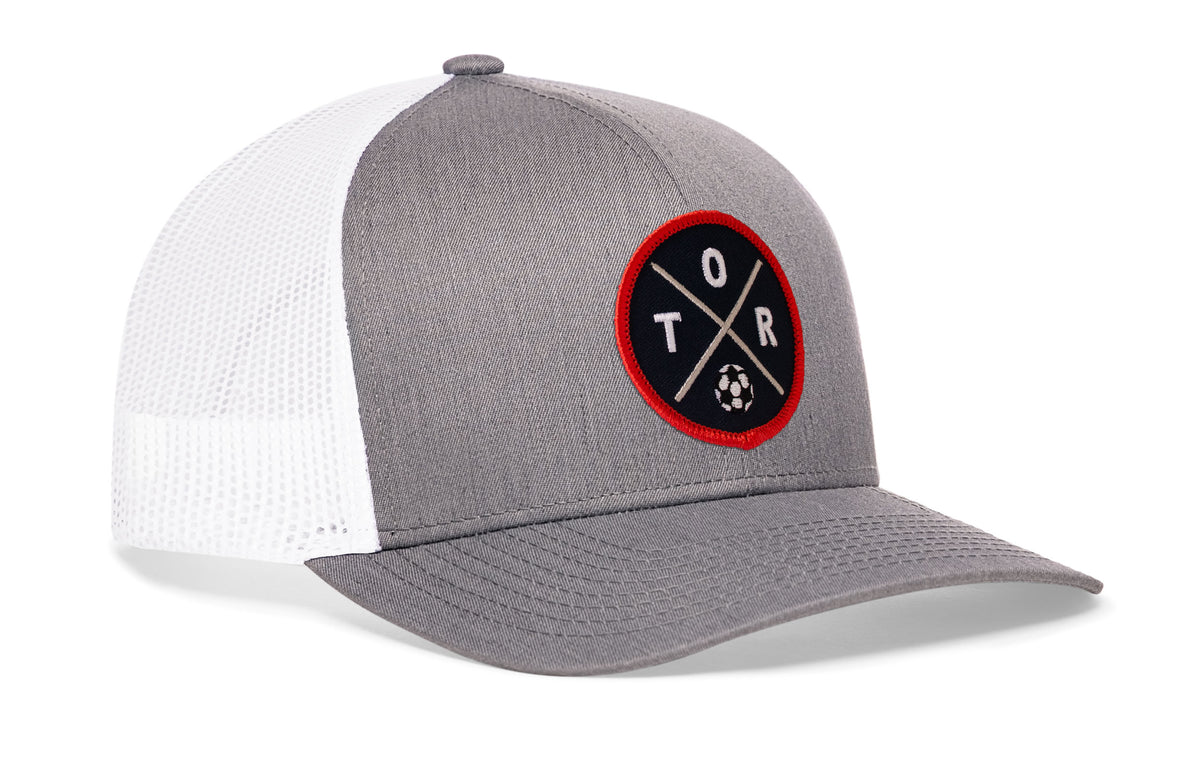 gray-white trucker hat viewed from the right side, showcasing a bold red and black embroidered X patch prominently displayed on the front