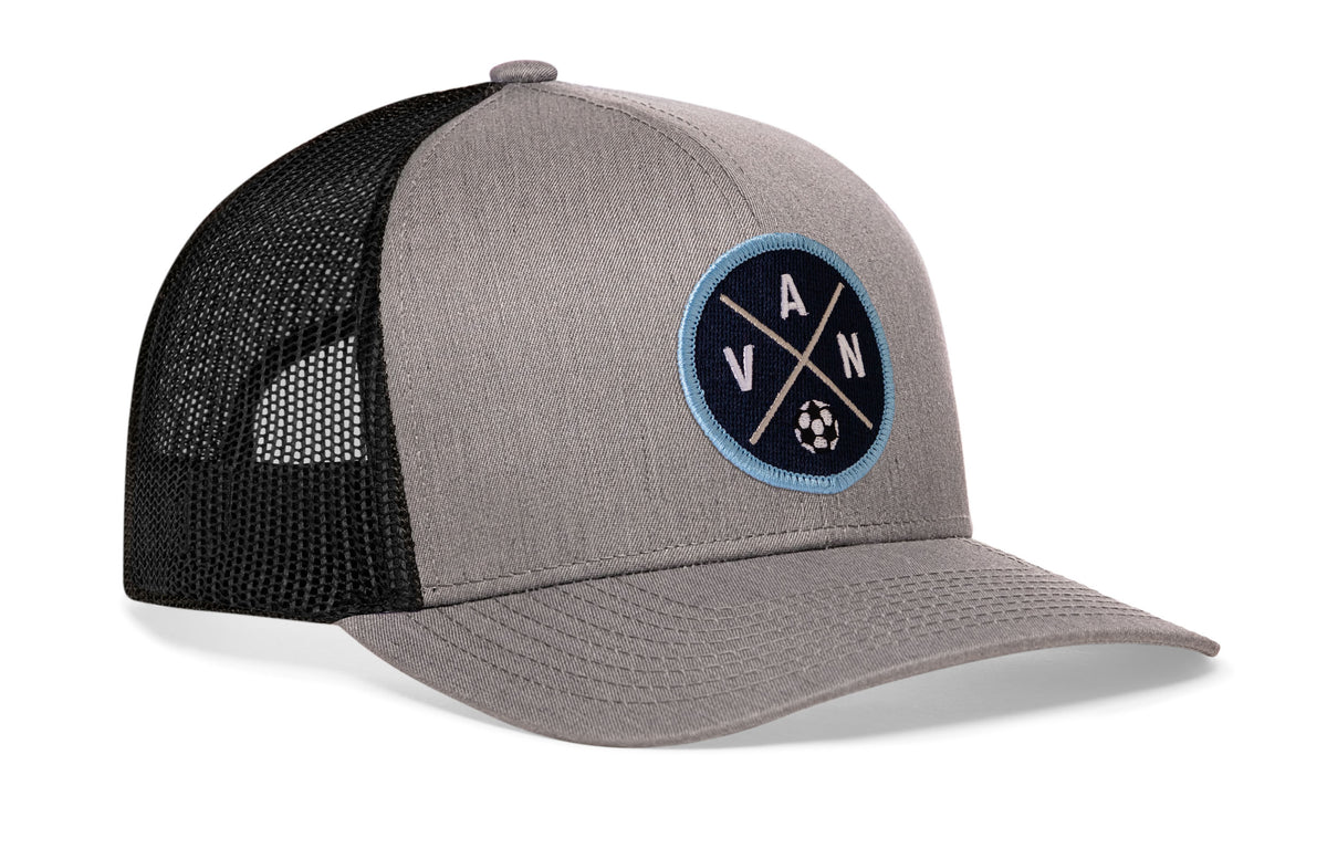 gray-black trucker hat viewed from the right side, showcasing a bold blue and white embroidered X patch prominently displayed on the front