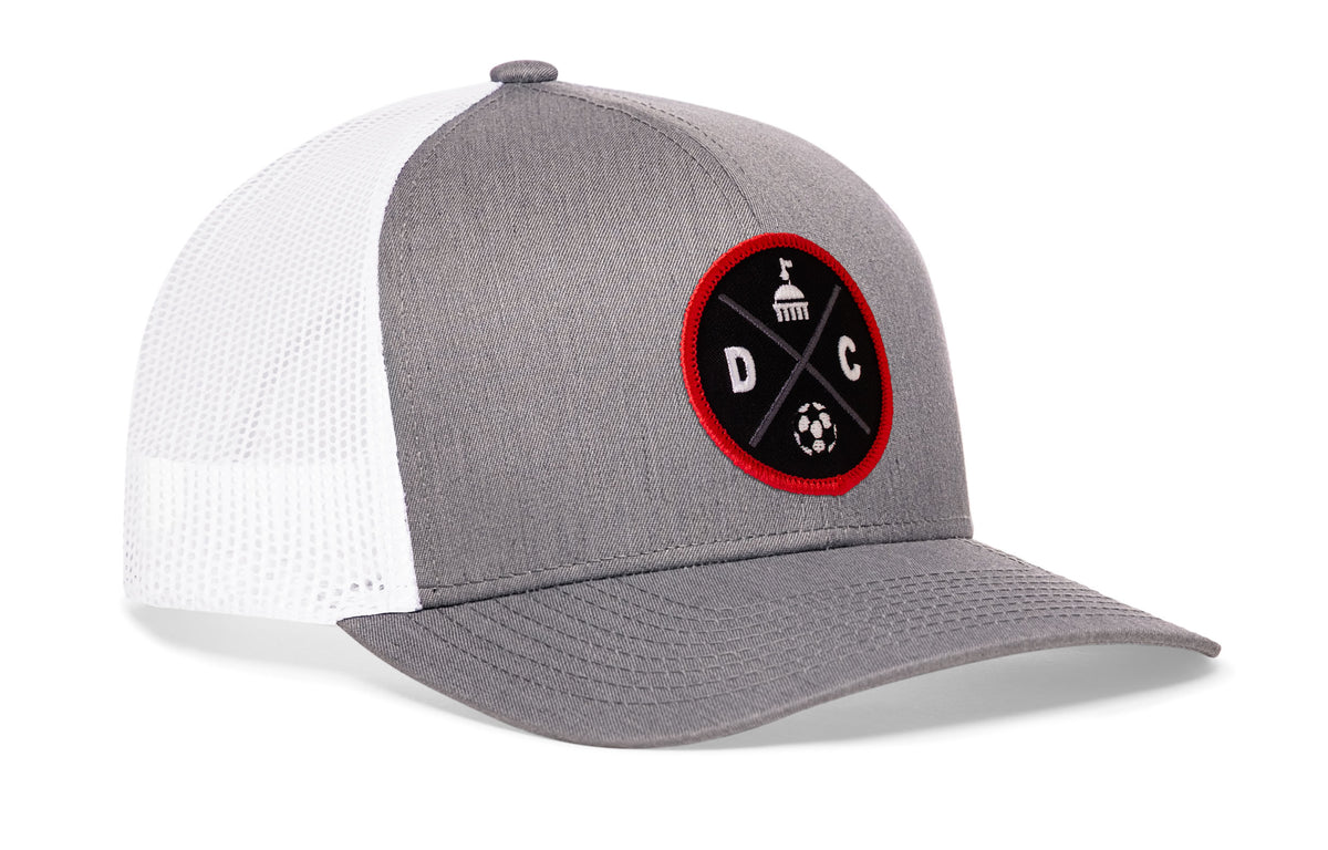 gray-white trucker hat viewed from the right side, showcasing a bold black and red embroidered X patch prominently displayed on the front