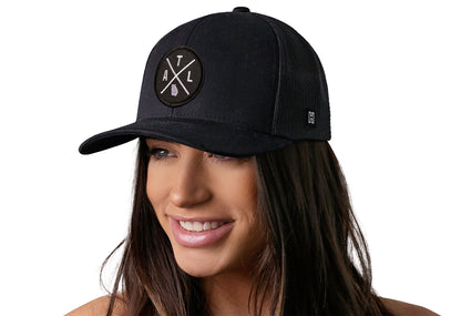 Black trucker hat worn by a female model viewed from the left side and features a Atlanta ATL X Embroidered Circle Patch displayed on the front panel. The hat has a classic mesh back design and a curved structured bill