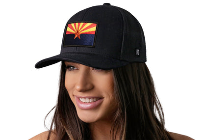 Black trucker hat worn by a female model viewed from the left side and features a Arizona Flag Embroidered Rectangle Patch displayed on the front panel. The hat has a classic mesh back design and a curved structured bill