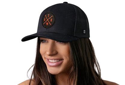 Black trucker hat worn by a female model viewed from the left side and features a Austin (Orange) ATX Embroidered X Patch displayed on the front panel. The hat has a classic mesh back design and a curved structured bill