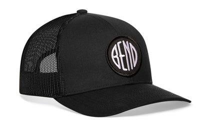 Black trucker hat viewed from the right side, showcasing a Bend Embroidered Circle Patch prominently displayed on the front