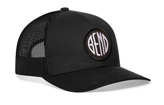 Black trucker hat viewed from the right side, showcasing a Bend Embroidered Circle Patch prominently displayed on the front