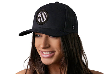 Black trucker hat worn by a female model viewed from the left side and features a Bend Embroidered Circle Patch displayed on the front panel. The hat has a classic mesh back design and a curved structured bill