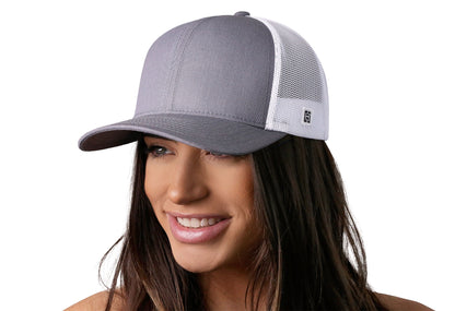 Gray-White trucker hat worn by a female model viewed from the left side and features a n/a, Blank Gray White Hat displayed on the front panel. The hat has a classic mesh back design and a curved structured bill