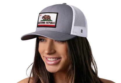 Gray-White trucker hat worn by a female model viewed from the left side and features a California Flag Embroidered Rectangle Patch displayed on the front panel. The hat has a classic mesh back design and a curved structured bill