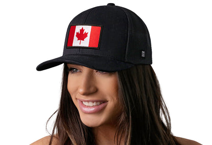 Black trucker hat worn by a female model viewed from the left side and features a Canadian Flag Embroidered Rectangle Patch displayed on the front panel. The hat has a classic mesh back design and a curved structured bill