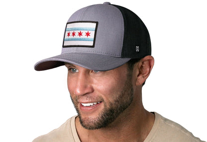 Gray-Black trucker hat worn by a male model viewed from the left side and features a Chicago Flag Embroidered Rectangle Patch displayed on the front panel. The hat has a classic mesh back design and a curved structured bill