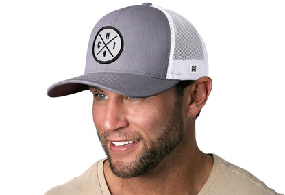 Gray-White trucker hat worn by a male model viewed from the left side and features a Chicago CHI X Embroidered White Circle Patch displayed on the front panel. The hat has a classic mesh back design and a curved structured bill