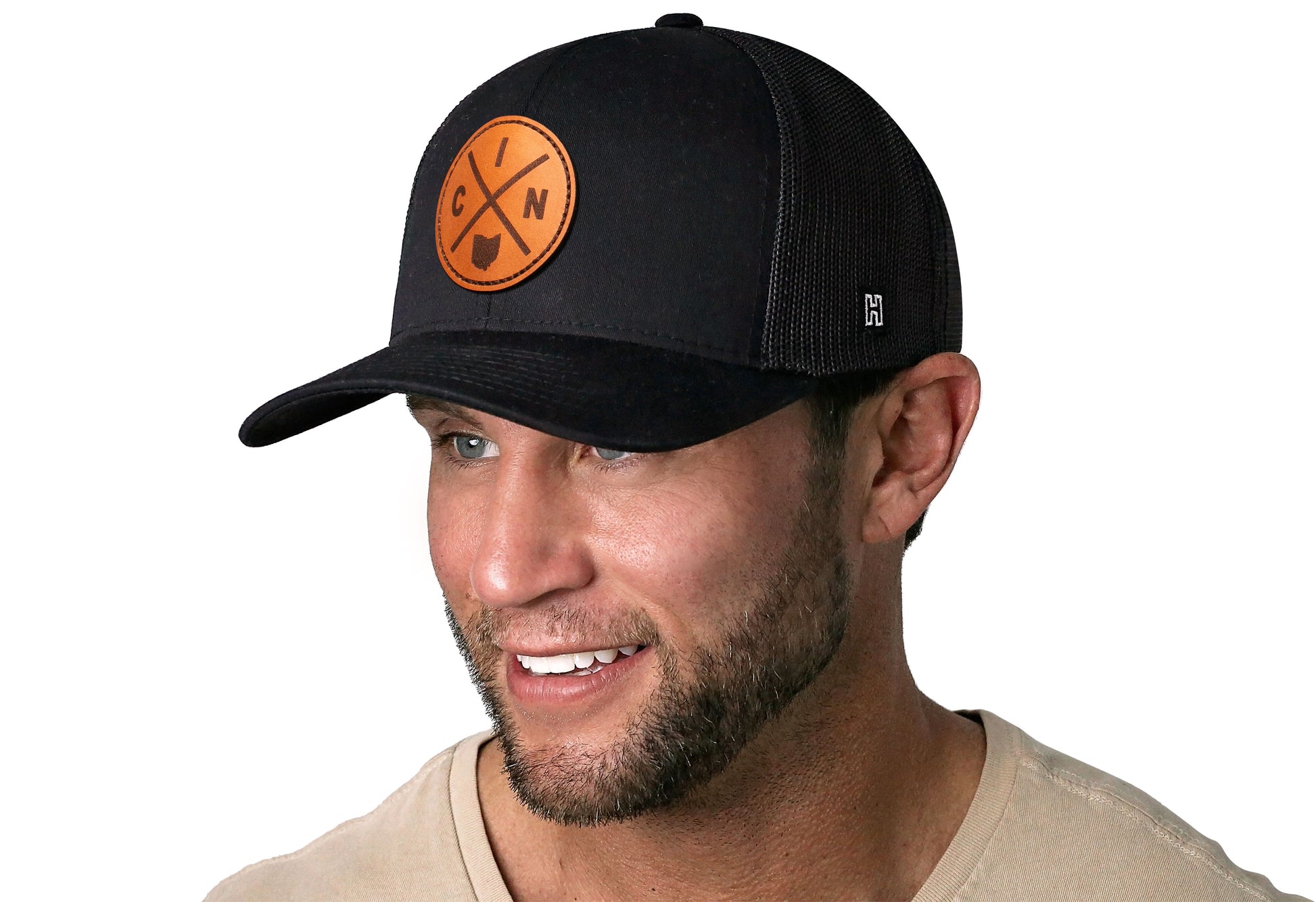 Black trucker hat worn by a male model viewed from the left side and features a Cincinnati CIN X Leather Circle Patch displayed on the front panel. The hat has a classic mesh back design and a curved structured bill
