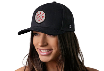 Black trucker hat worn by a female model viewed from the left side and features a Columbus CBUS X Gray, Red Embroidered Circle Patch displayed on the front panel. The hat has a classic mesh back design and a curved structured bill