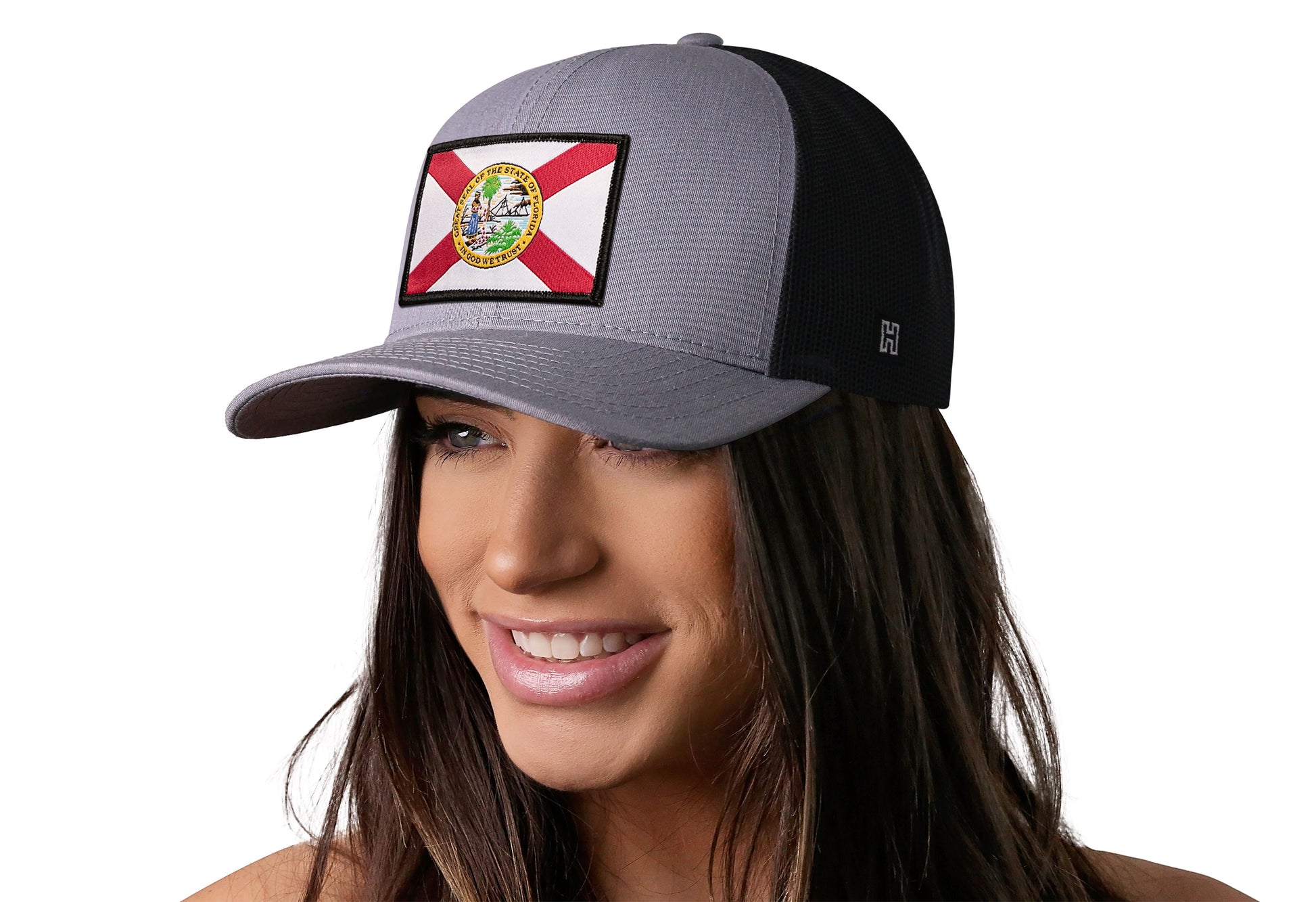Gray-Black trucker hat worn by a female model viewed from the left side and features a Florida Flag Woven Retangle Patch displayed on the front panel. The hat has a classic mesh back design and a curved structured bill