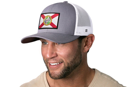Gray-White trucker hat worn by a male model viewed from the left side and features a Florida Flag Woven Retangle Patch displayed on the front panel. The hat has a classic mesh back design and a curved structured bill