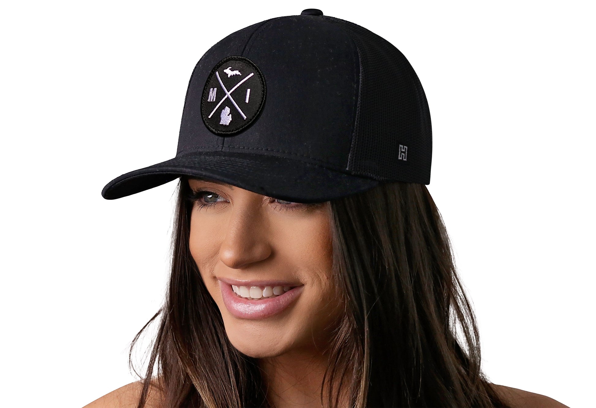 Black trucker hat worn by a female model viewed from the left side and features a Michigan MI X Embroidered Circle Patch displayed on the front panel. The hat has a classic mesh back design and a curved structured bill