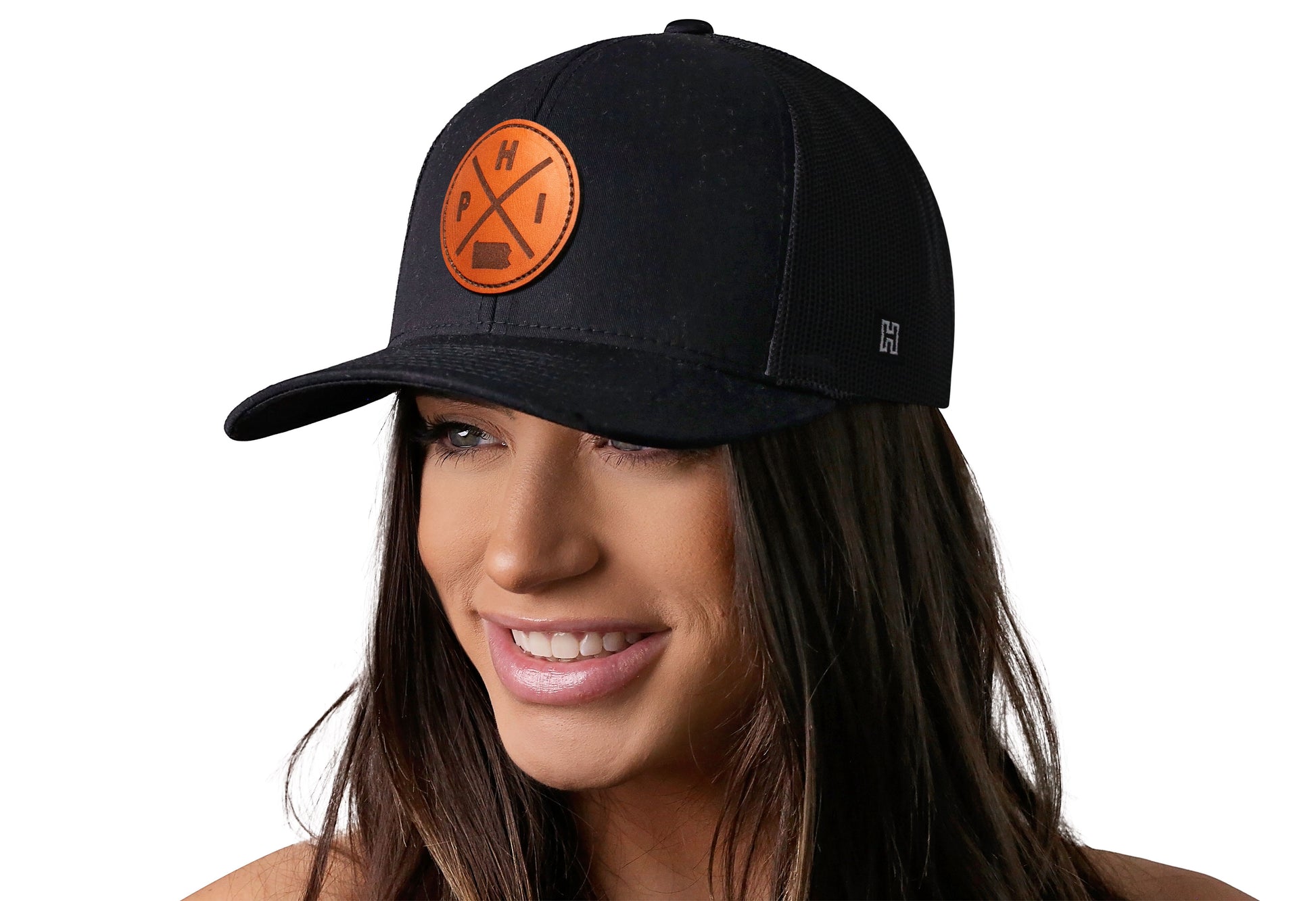 Black trucker hat worn by a female model viewed from the left side and features a Philadelphia PHI X Leather Circle Patch displayed on the front panel. The hat has a classic mesh back design and a curved structured bill