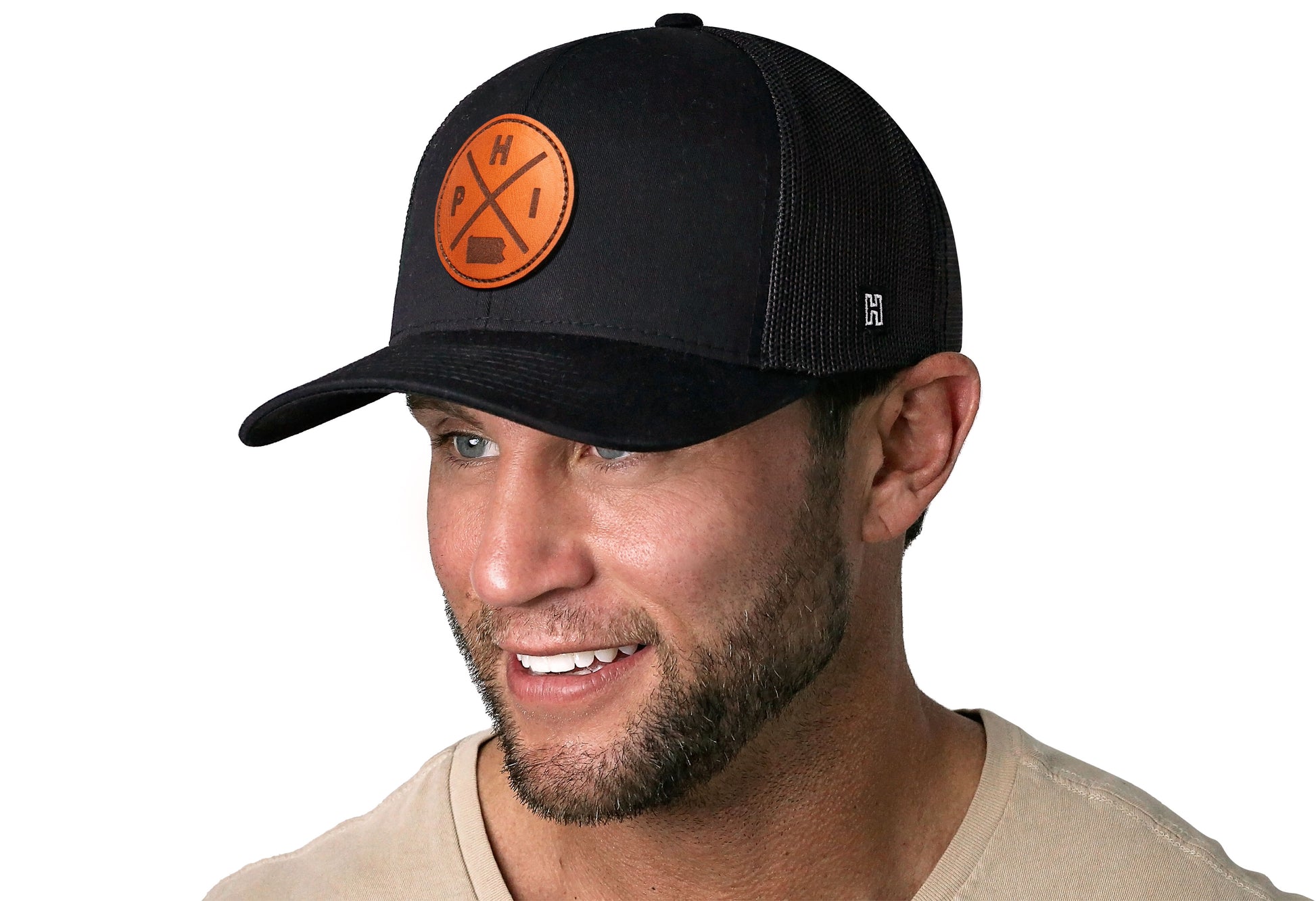 Black trucker hat worn by a male model viewed from the left side and features a Philadelphia PHI X Leather Circle Patch displayed on the front panel. The hat has a classic mesh back design and a curved structured bill