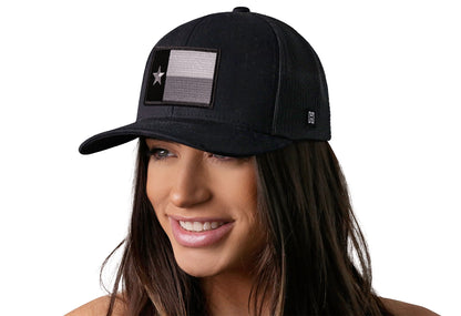 Black trucker hat worn by a female model viewed from the left side and features a Texas TX Tactical Embroidered Flag Patch displayed on the front panel. The hat has a classic mesh back design and a curved structured bill