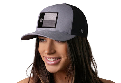 Gray-Black trucker hat worn by a female model viewed from the left side and features a Texas Gray-Black TX Tactical Embroidered Flag Patch displayed on the front panel. The hat has a classic mesh back design and a curved structured bill