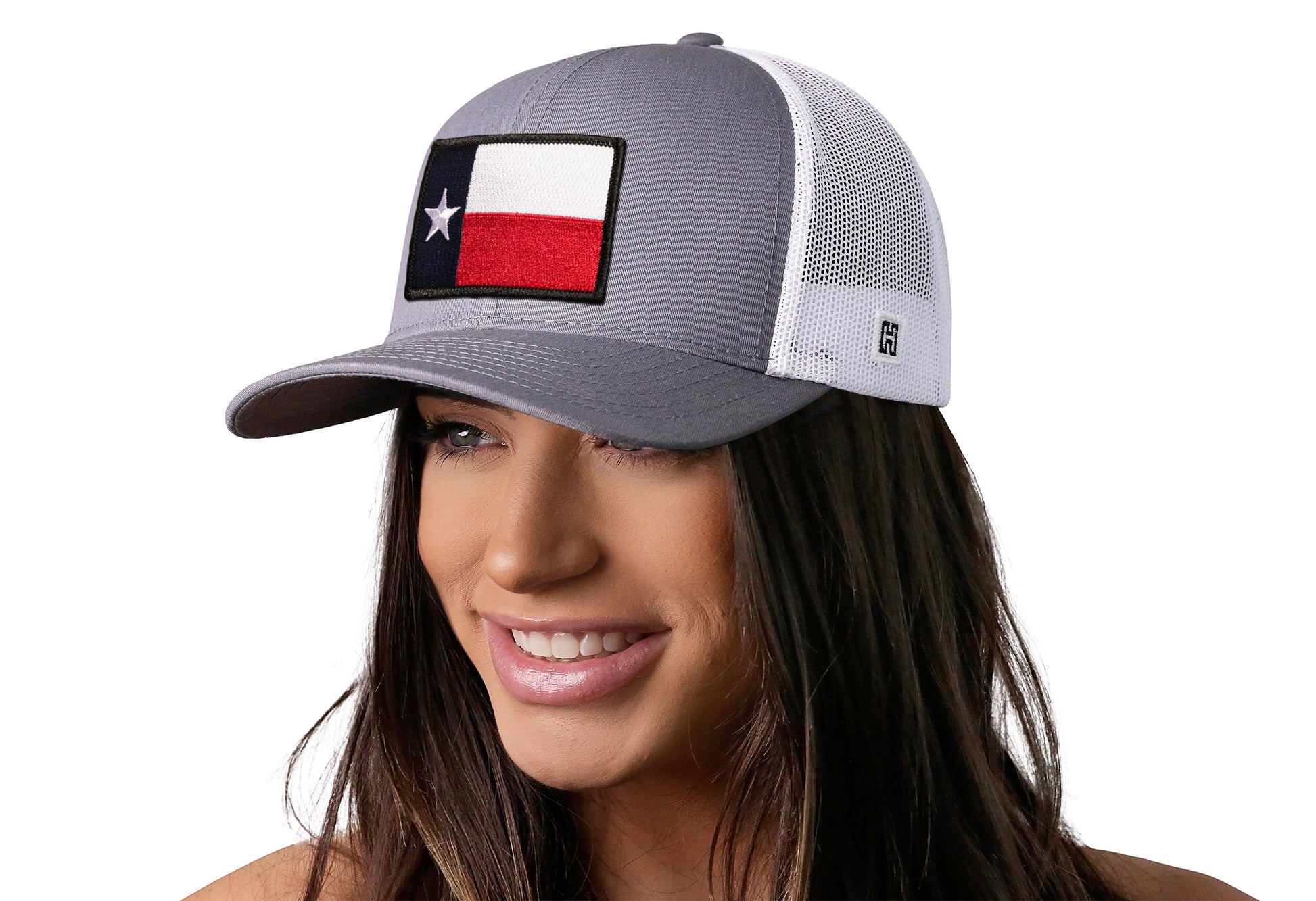 Gray-White trucker hat worn by a female model viewed from the left side and features a Texas TX Embroidered Flag Patch displayed on the front panel. The hat has a classic mesh back design and a curved structured bill